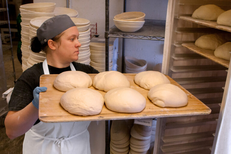 A CRUST baker carrying a board of shaped loaves past stacks of proofing baskets