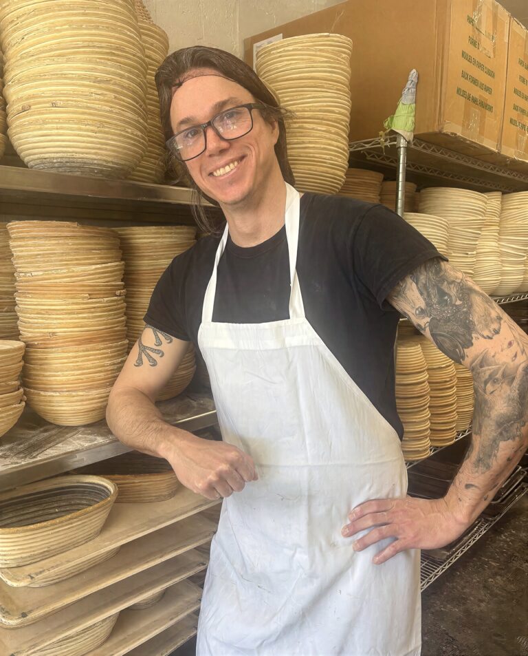Cameron Alesso of the CRUST Bread Team, smiling beside shelves of bread proofing baskets